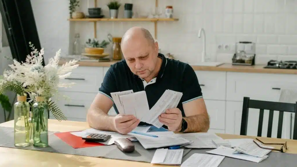 A man sits at a kitchen table covered in papers and bills, looking at documents.