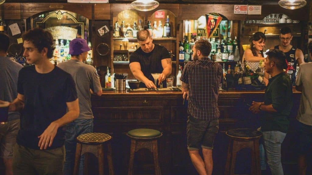 A bartender prepares a drink behind a crowded bar while several people stand around talking.