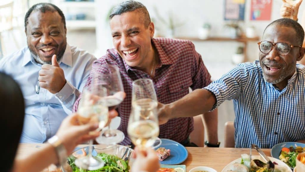 Three men sit at a dining table laughing while clinking wine glasses during a meal.