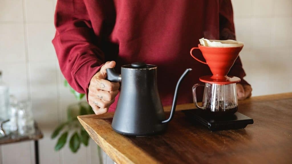 A person in a red sweatshirt holds a black kettle while preparing pour-over coffee.