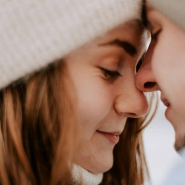 A close-up shot of a couple wearing beanies, leaning in with their foreheads and noses touching.