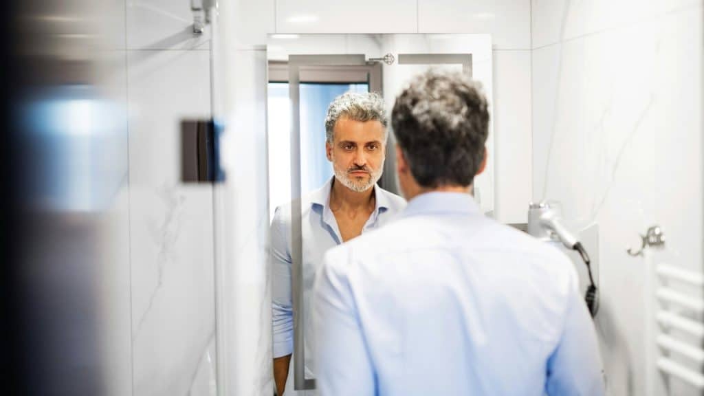 A man with grey hair looks at his own reflection in a brightly lit bathroom mirror.