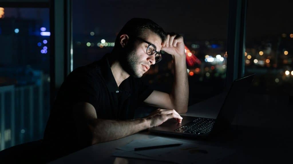 A man wearing glasses sits at a desk in a dark room looking at a laptop.