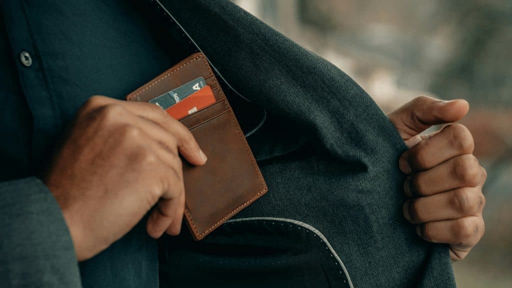 A person in a grey suit jacket pulls a brown leather cardholder from an inner pocket.