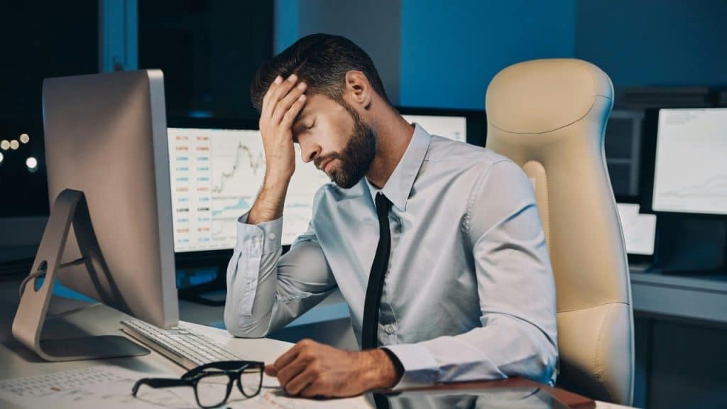 A man in a shirt and tie sits at a desk, holding his head in distress.