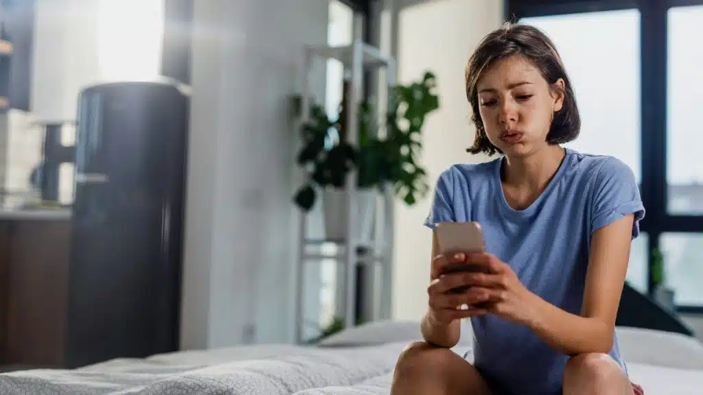A woman in a blue shirt sits on a bed, looking down at her phone.