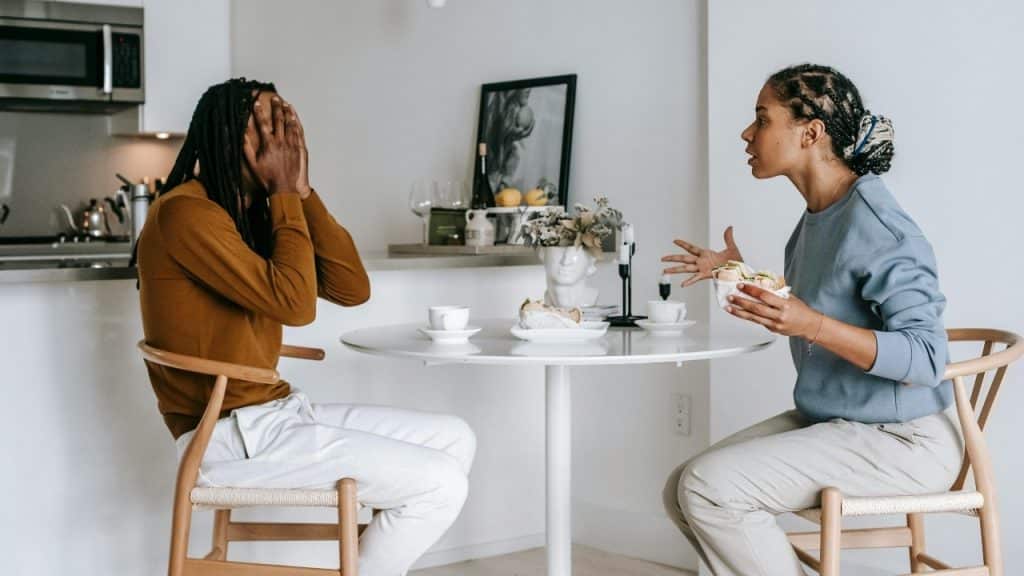 A man covers his face while a woman gestures and speaks at a dining table.