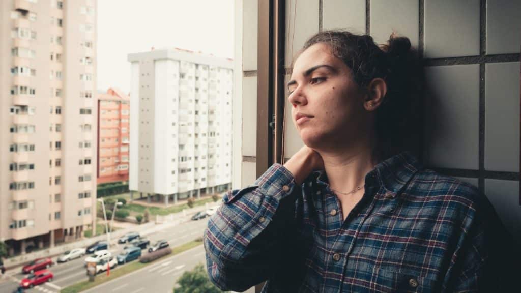 A woman in a plaid shirt stands by a window, looking out at city buildings.