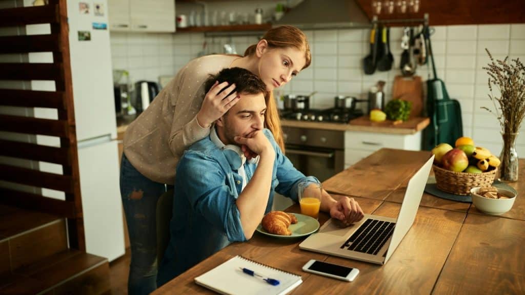 A woman rests her hand on a man’s head while they look at a laptop.