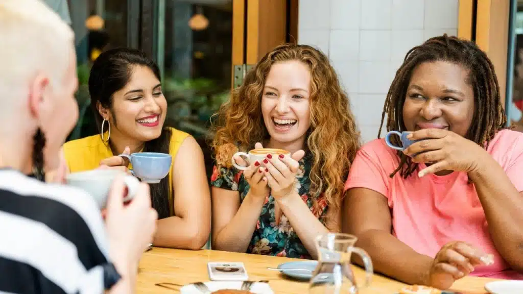 Four women sit at a wooden table together, smiling and drinking from colorful coffee mugs.