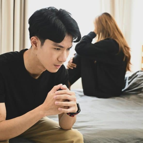 A man sits on a bed with hands clasped while a woman sits facing away.