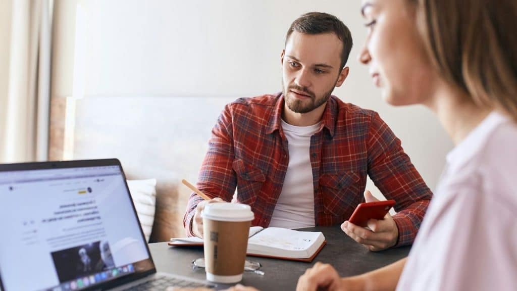 A man with a notebook and phone looks at a woman sitting across from a laptop.