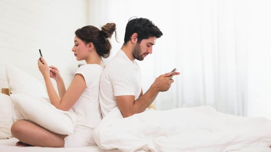A man and woman sit back to back on a bed while looking at their phones.