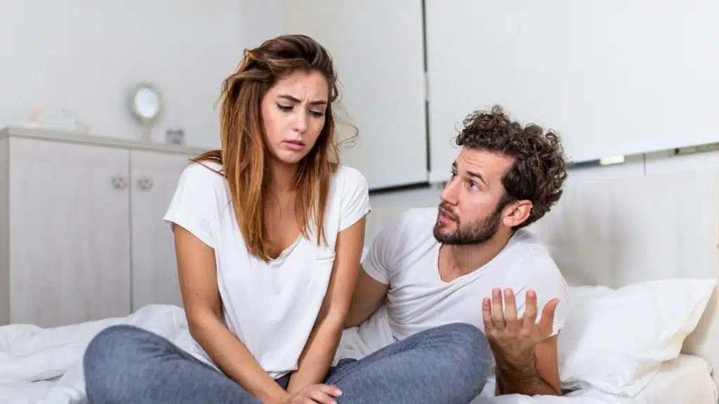 A woman sits on a bed looking down while a man beside her gestures mid-speech.