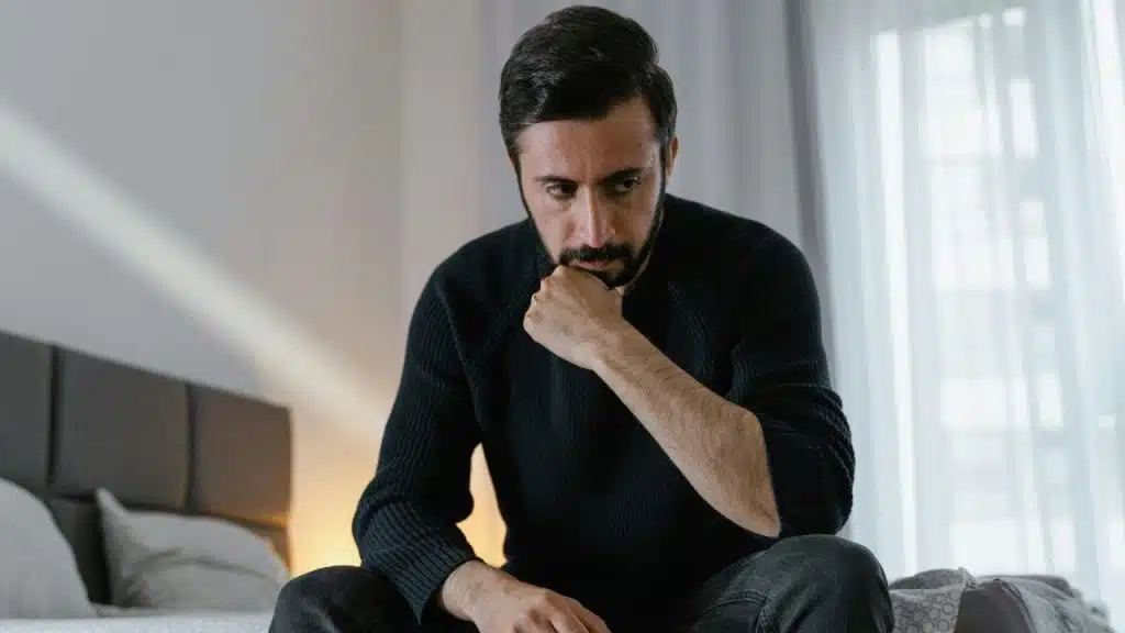 A bearded man in a black sweater sits on a bed, resting his chin on his fist.