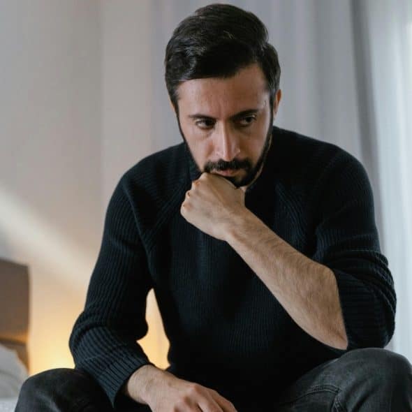 A bearded man in a black sweater sits on a bed, resting his chin on his fist.