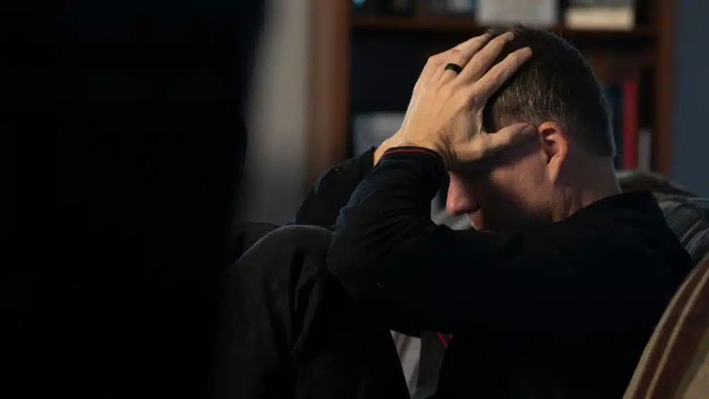 A man sits in a dark room with his hands on his head, looking downward in distress.