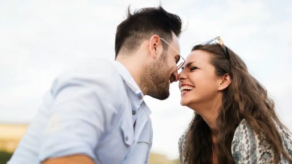 A man and woman face each other closely, smiling and laughing with their foreheads almost touching.
