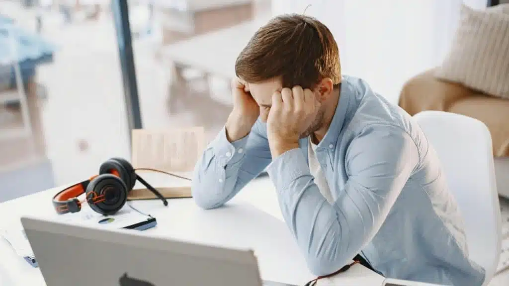 A man sits at a white desk with his head in his hands near a laptop.