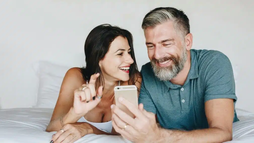 A woman and a gray-haired man lie on a bed together, smiling at a smartphone screen.