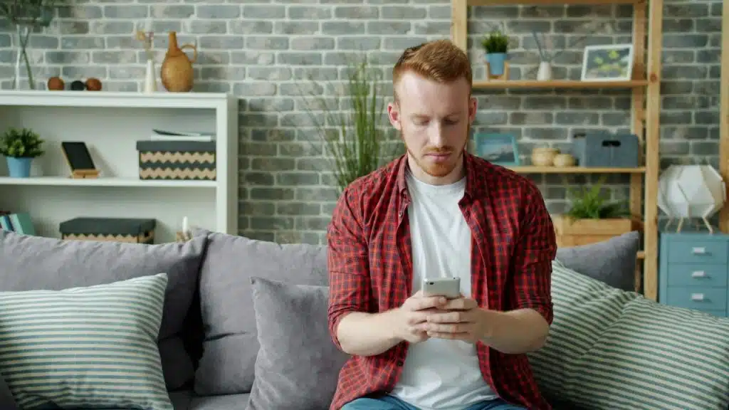 A man in a red plaid shirt sits on a gray sofa while looking at a smartphone.