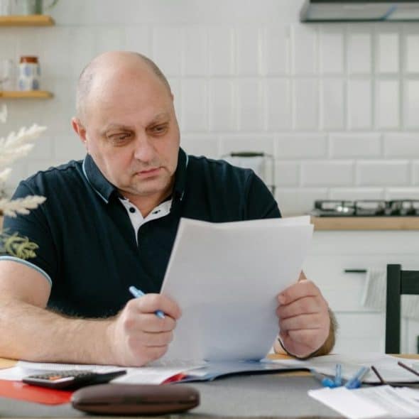A man in a black polo shirt sits at a table while reviewing paper documents.