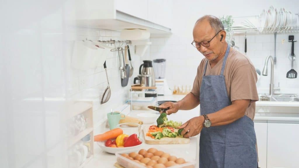 An older man wearing a blue apron chops vegetables in a bright, modern white kitchen.