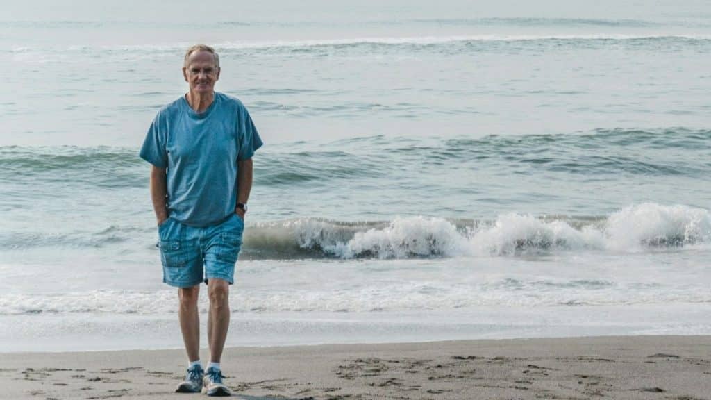 A man in a blue shirt and shorts walks along a sandy beach near waves.