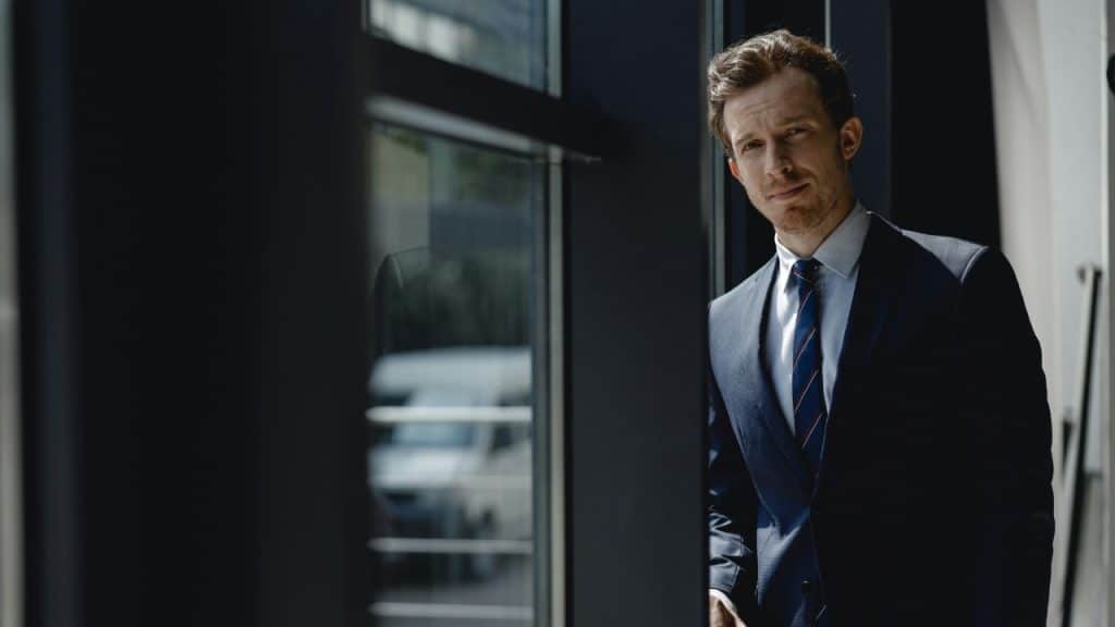 A man in a dark suit and striped tie stands next to a large window.