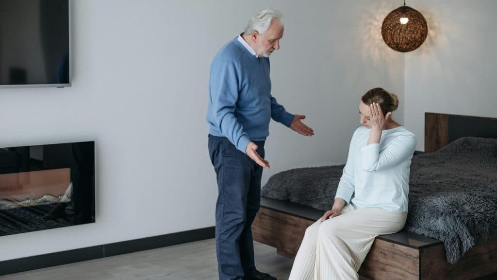 An older man gestures toward a woman sitting on a bed with her hand to her head.
