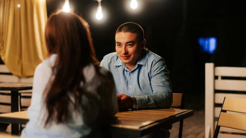 A man in a blue shirt sits at an outdoor table looking at a woman.