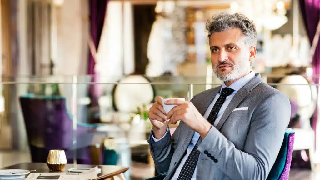 A man with grey hair and a suit holding a small white cup in a cafe.
