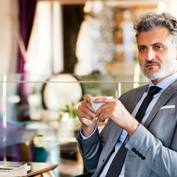A man with grey hair and a suit holding a small white cup in a cafe.