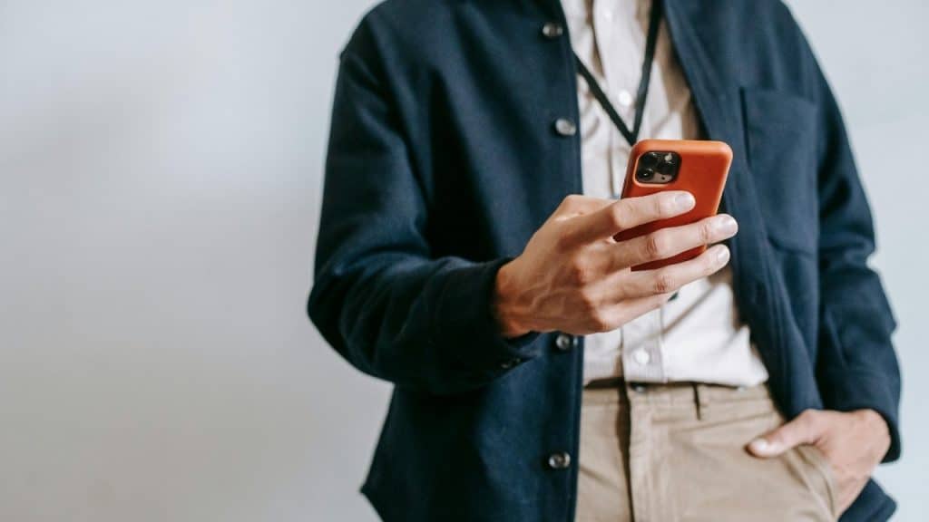 A person in a dark jacket holds an orange smartphone with one hand in their pocket.