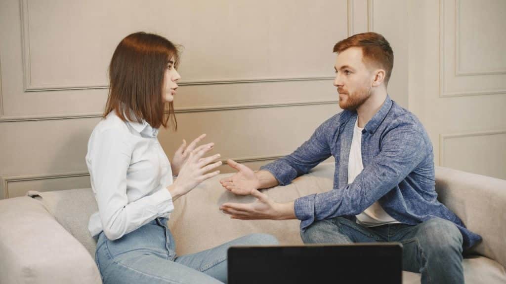 A man and woman sit on a sofa gesturing with their hands while talking.