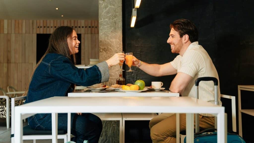 A man and woman sit at a white table clinking juice glasses together while smiling.