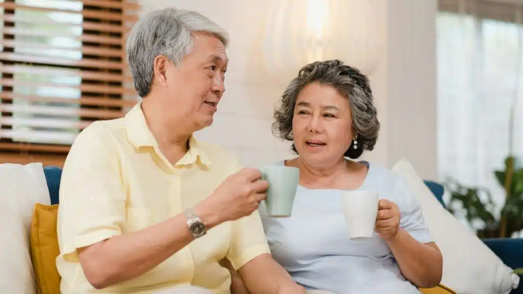 An older man and woman sit on a couch, holding mugs and talking to each other.