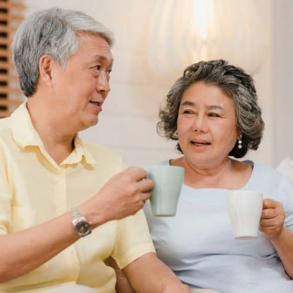 An older man and woman sit on a couch, holding mugs and talking to each other.