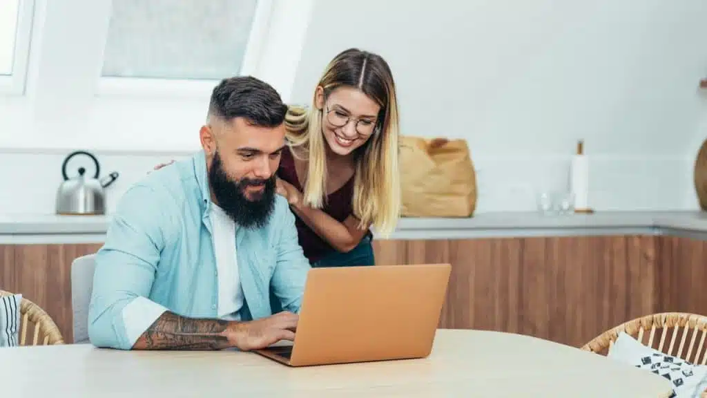 A man with a beard uses a laptop while a woman stands behind him and smiles.