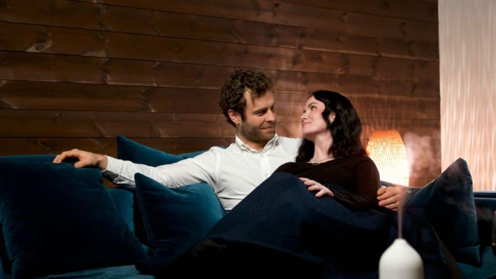 A man and woman sit together on a blue velvet sofa in a dimly lit room.