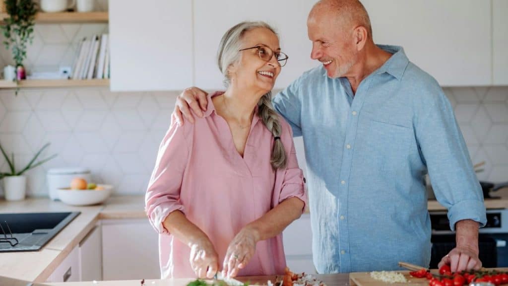 An older man smiles at a woman as she chops vegetables in a bright, modern kitchen.