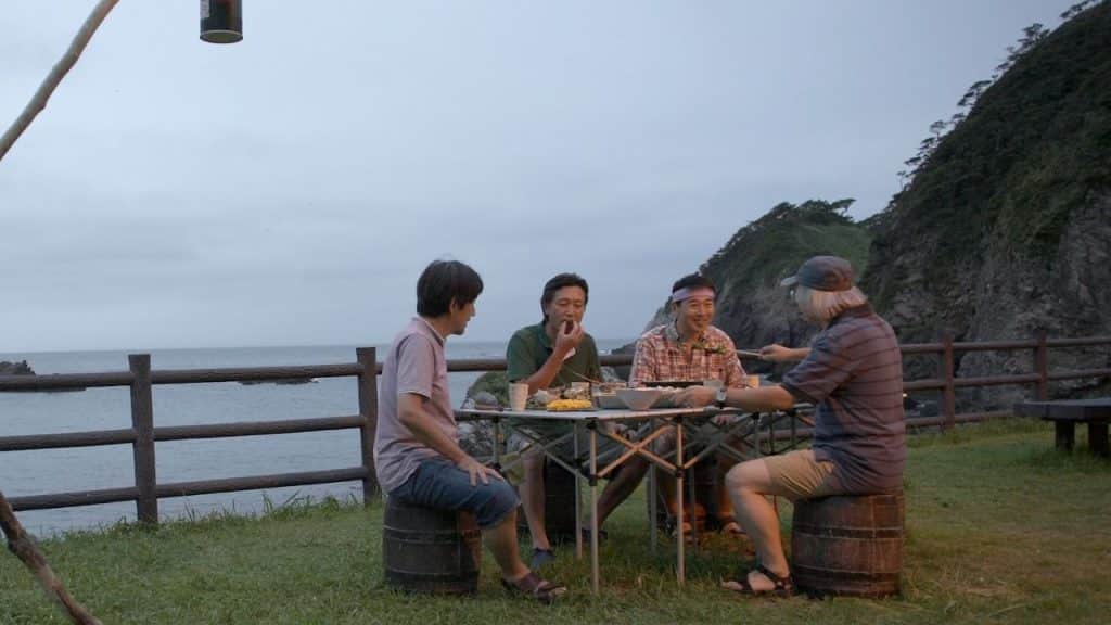 Four men sit around a table outdoors near a fence with a view of the ocean.