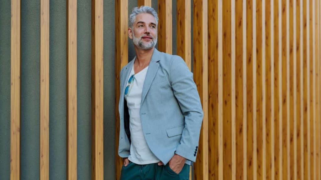 A man with grey hair leans against a wall made of vertical wooden slats outdoors.
