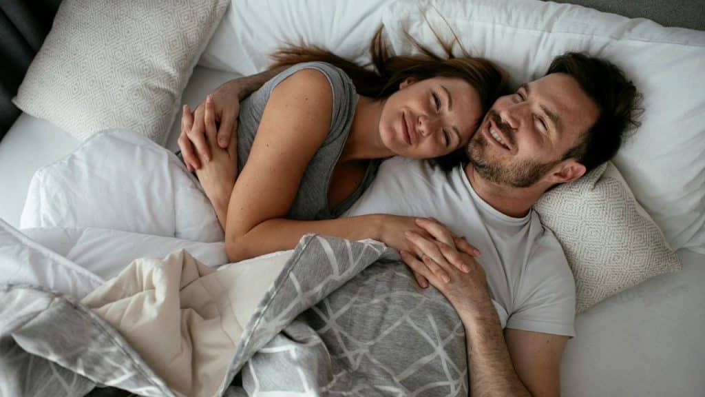 A man and woman lie in bed under a grey blanket, smiling and holding hands together.