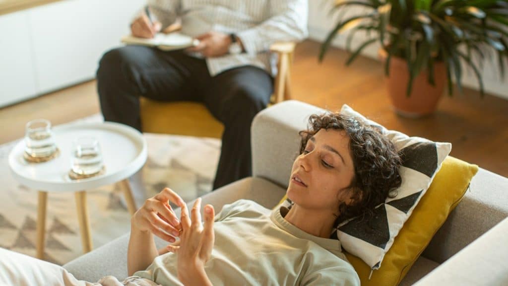A mentally depressed woman looking at her own hands while lying on a couch and a man is noting down something while sitting next to her.