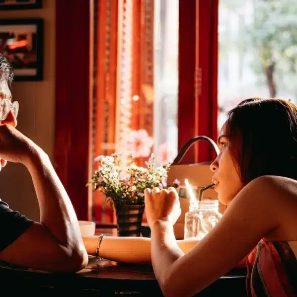 Two people sit across from each other at a small café table by a window, engaged in conversation.