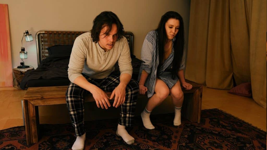 Two people sit on the edge of a bed in a dimly lit bedroom, both looking down and appearing tense or worried.