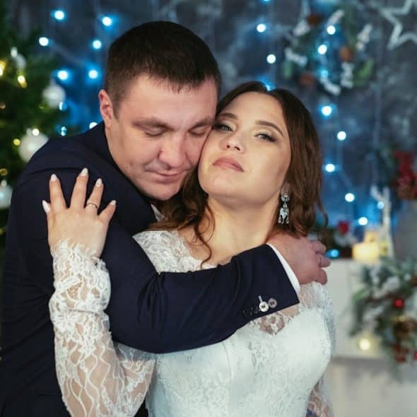 The man hugs the woman from behind with his eyes closed, while she looks calmly toward the camera.