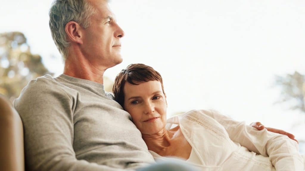 Mature couple relaxing together on a sofa, the woman resting her head on the man’s chest, sharing a calm, intimate moment.