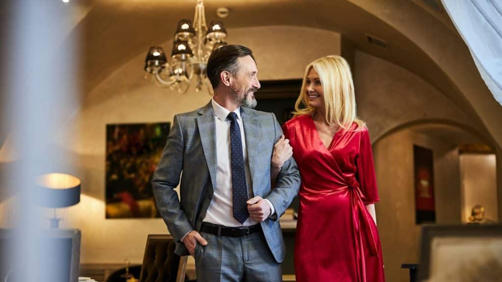 A cheerful blonde woman in red gown locking arms with an elegant bearded man while entering a fancy dining place.